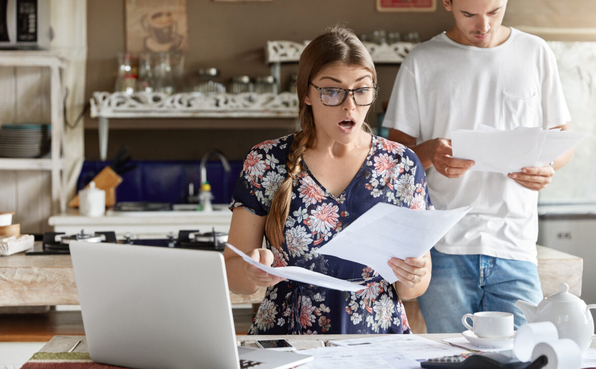 Young female looks with shocked expression into papers, recieve high taxes and had to pay much money for bills, sits against kitchen interior, her husband stands behinds, studies documents attentively